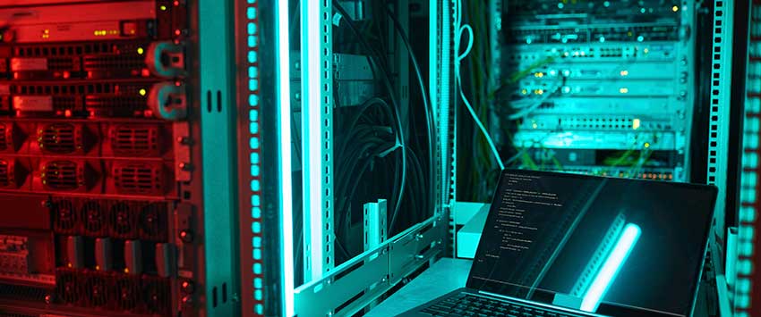 An image of a server room with racks filled with equipment illuminated by blue and red lights. A laptop is open in the foreground, displaying lines of code. This scene captures the technological aspect of modern data centers, emphasizing the critical role of infrastructure in digital communications and information security.