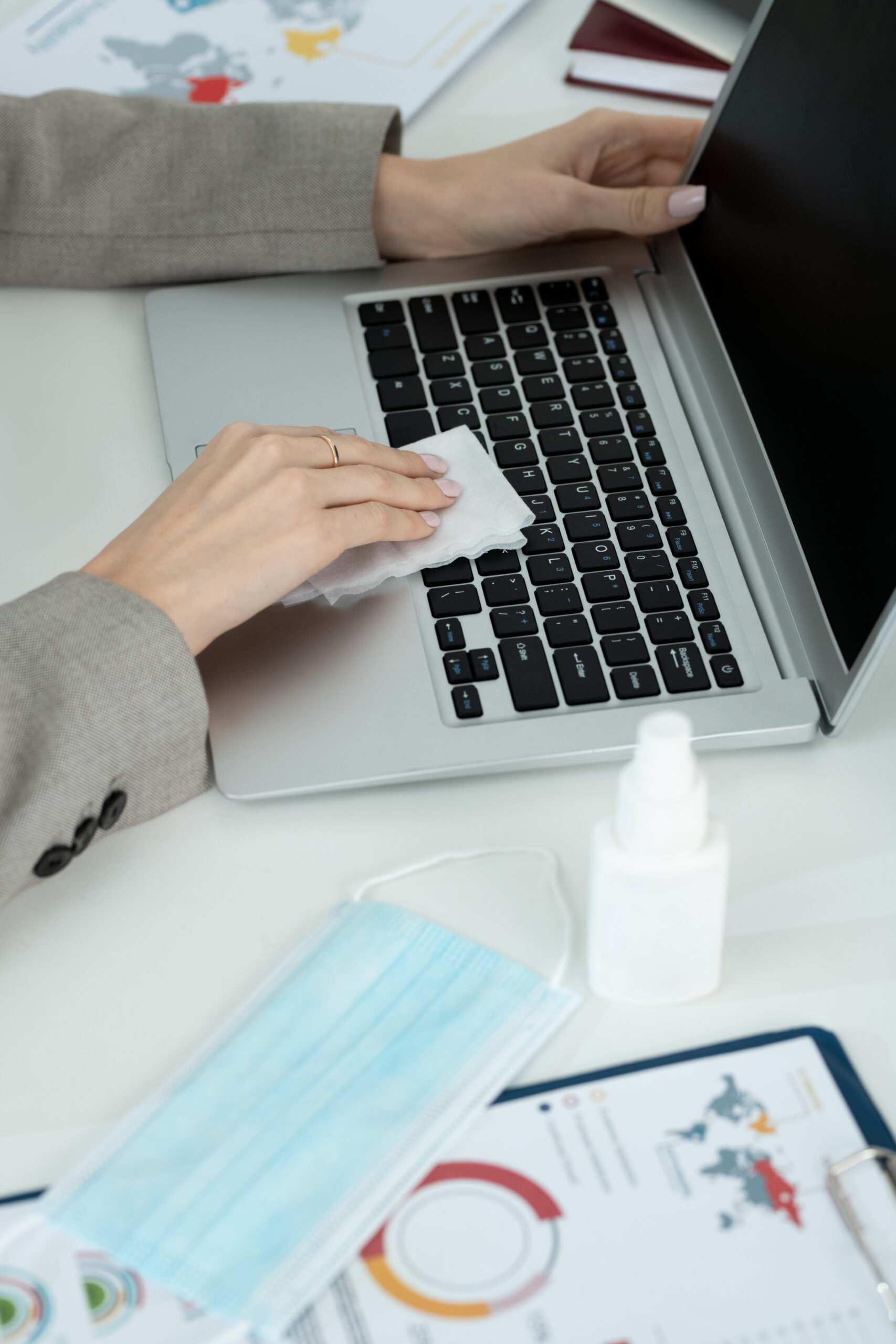 Hands of young contemporary businesswoman in grey formalwear wiping laptop keypad with sanitizer while sitting by workplace in office