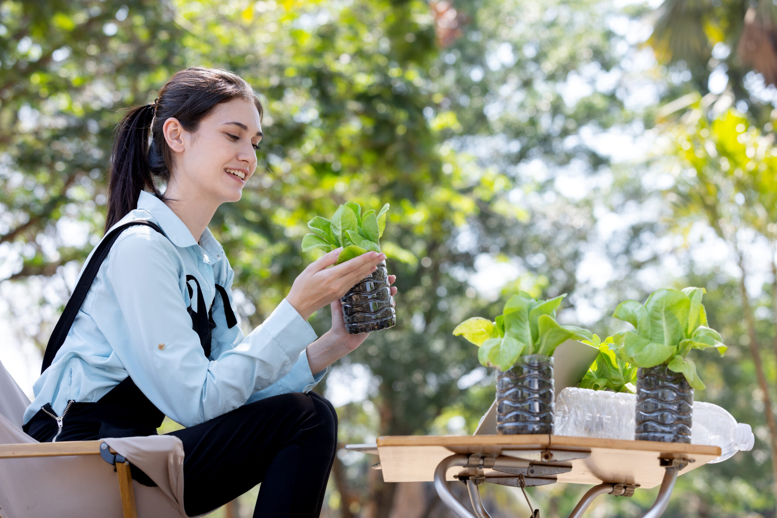 A young woman smiles as she examines a small, leafy plant in a recycled bottle planter. She's outdoors, with trees in the background, adding a lush green to the scene. The plant is one of several on a wooden cart, suggesting a mobile gardening project.