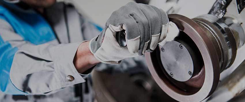 This image features a close-up view of a worker's hands, clad in protective gloves, engaged in the precise task of assembling or repairing industrial machinery. The worker's gray uniform suggests a professional setting, focused on heavy-duty mechanical work. The background is intentionally blurred to emphasize the detail and importance of the manual work being performed.