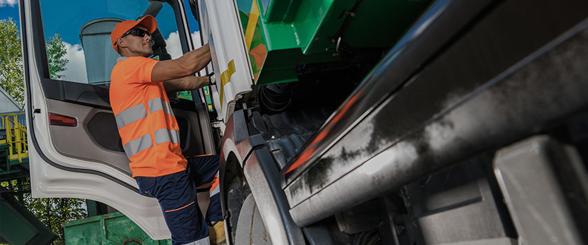 A worker in a reflective orange and blue uniform is seen stepping into a large green garbage truck.
