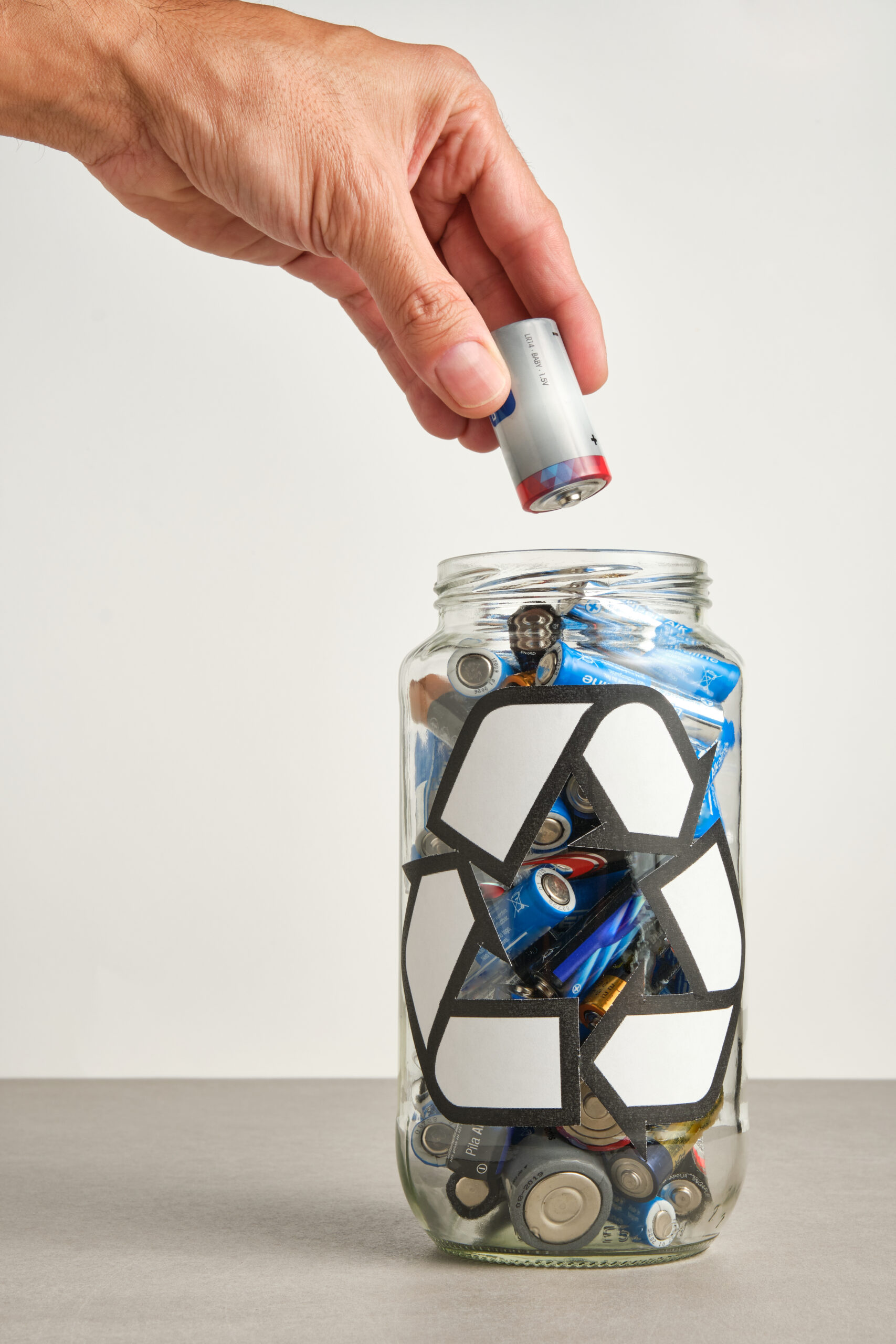 hand of a man pouring a used battery into a glass jar with the recycle symbol filled with used batteries on a neutral background