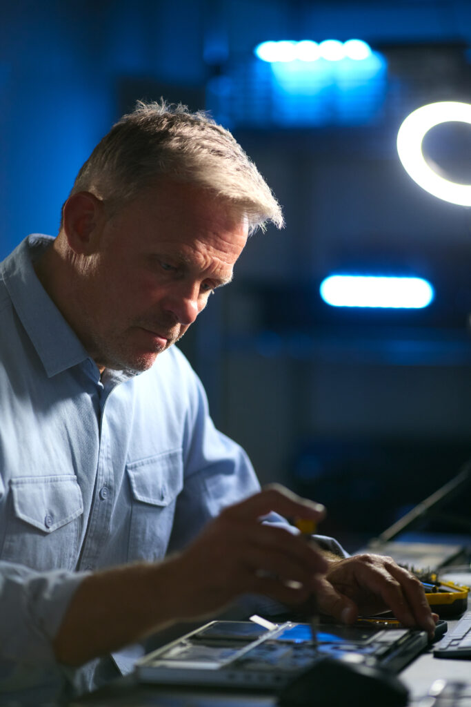 Mature Male Electronics Expert Repairing Laptop In Workshop With Low Key Lighting