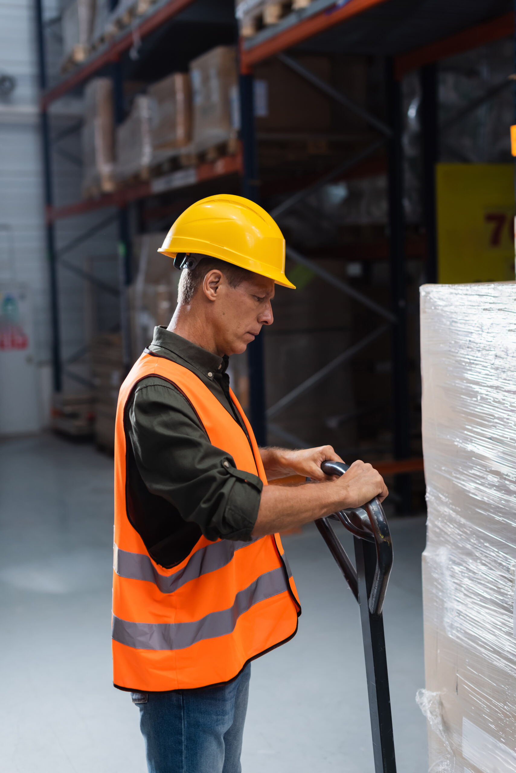 middle aged warehouse worker in hard hat and safety vest transporting pallet with a hand truck